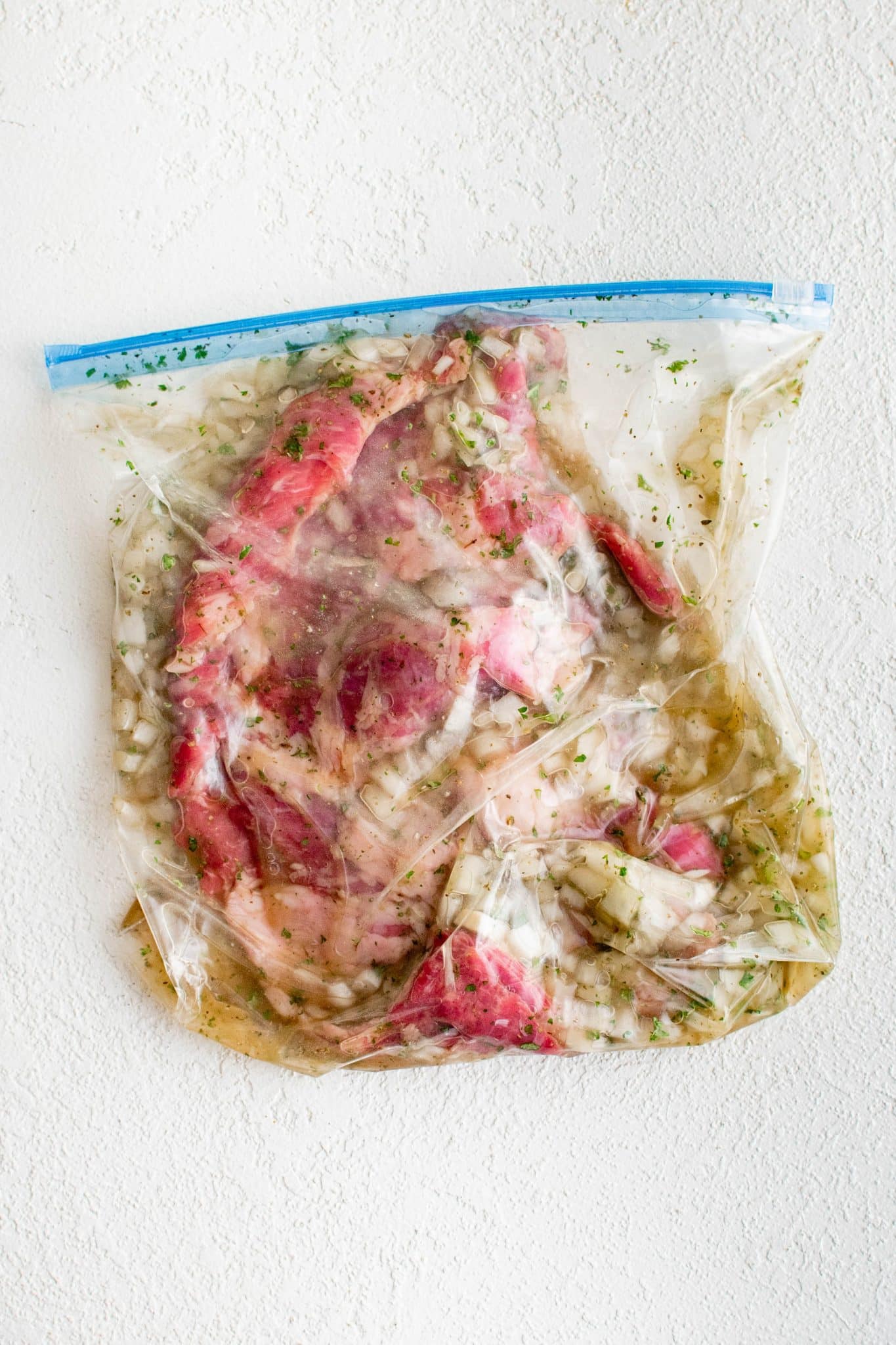 Overhead view of skirt steak sealed inside a clear plastic bag filled with marinade. The steak is fully submerged in a liquid mixture containing finely chopped onions, herbs, and seasonings, coating the meat on all sides.