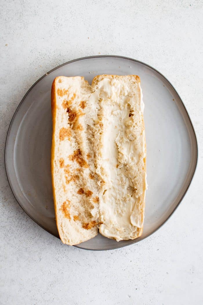 One French baguette on a plate cut in half lengthwise with mayonnaise spread on one half and jugo maggi drizzled on the other side.