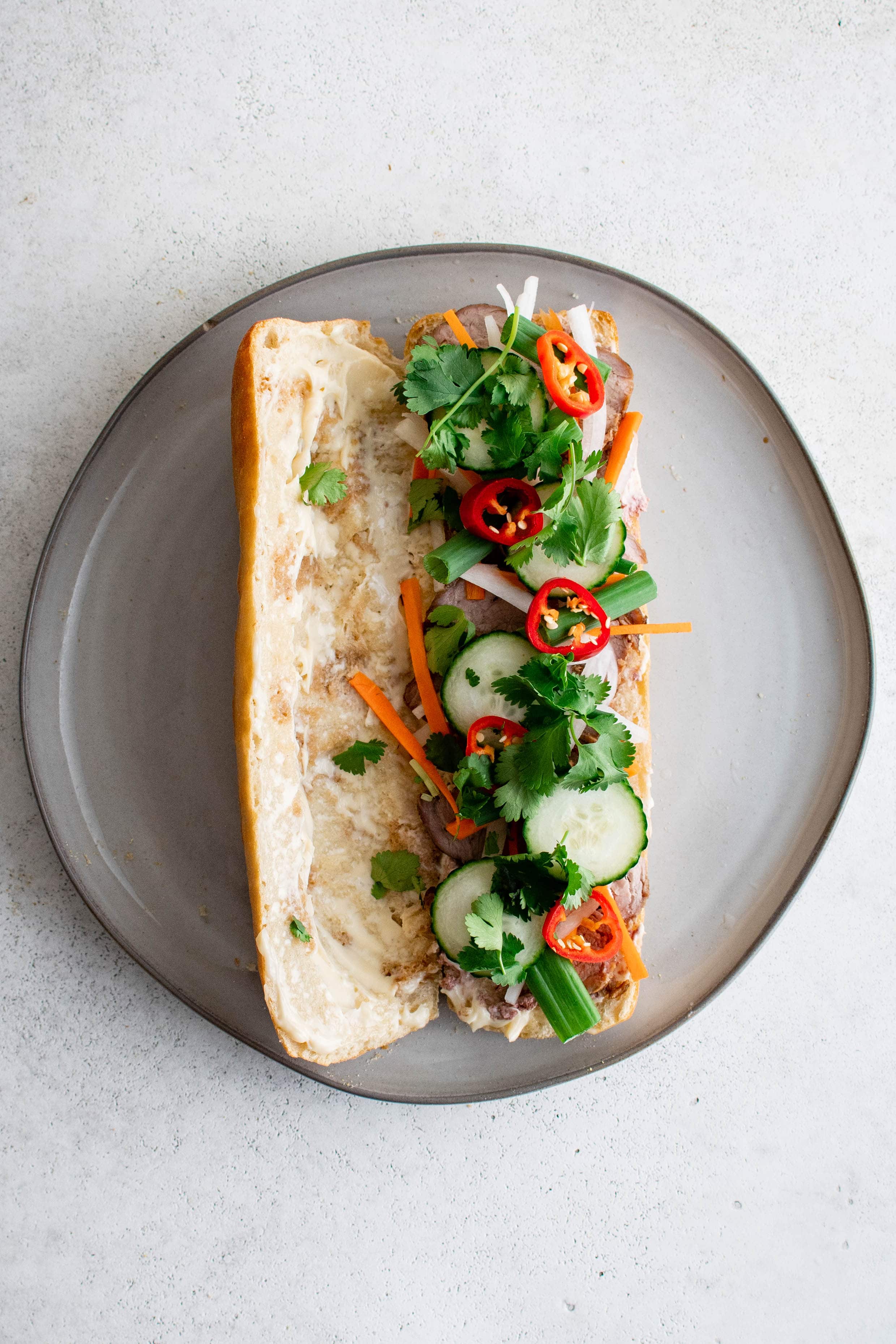 One French baguette on a plate cut in half lengthwise with one half topped with thinly sliced pork tenderloin, pickled radish and carrots, green onions, cucumber, and cilantro.