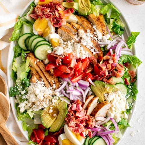 White serving plate with romaine lettuce topped with grilled chicken, bacon, hard boiled egg, tomatoes, feta cheese, cucumber, avocado, and red ion with red wine vinaigrette.