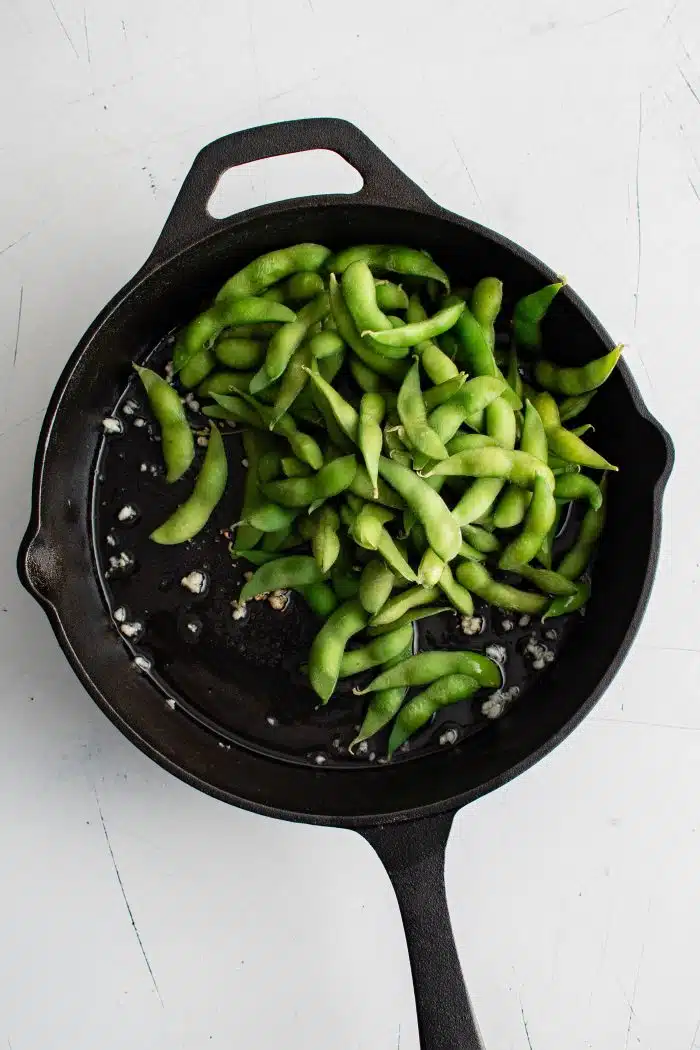 Thawed frozen edamame added to a large cast iron skillet with sauteed minced garlic.
