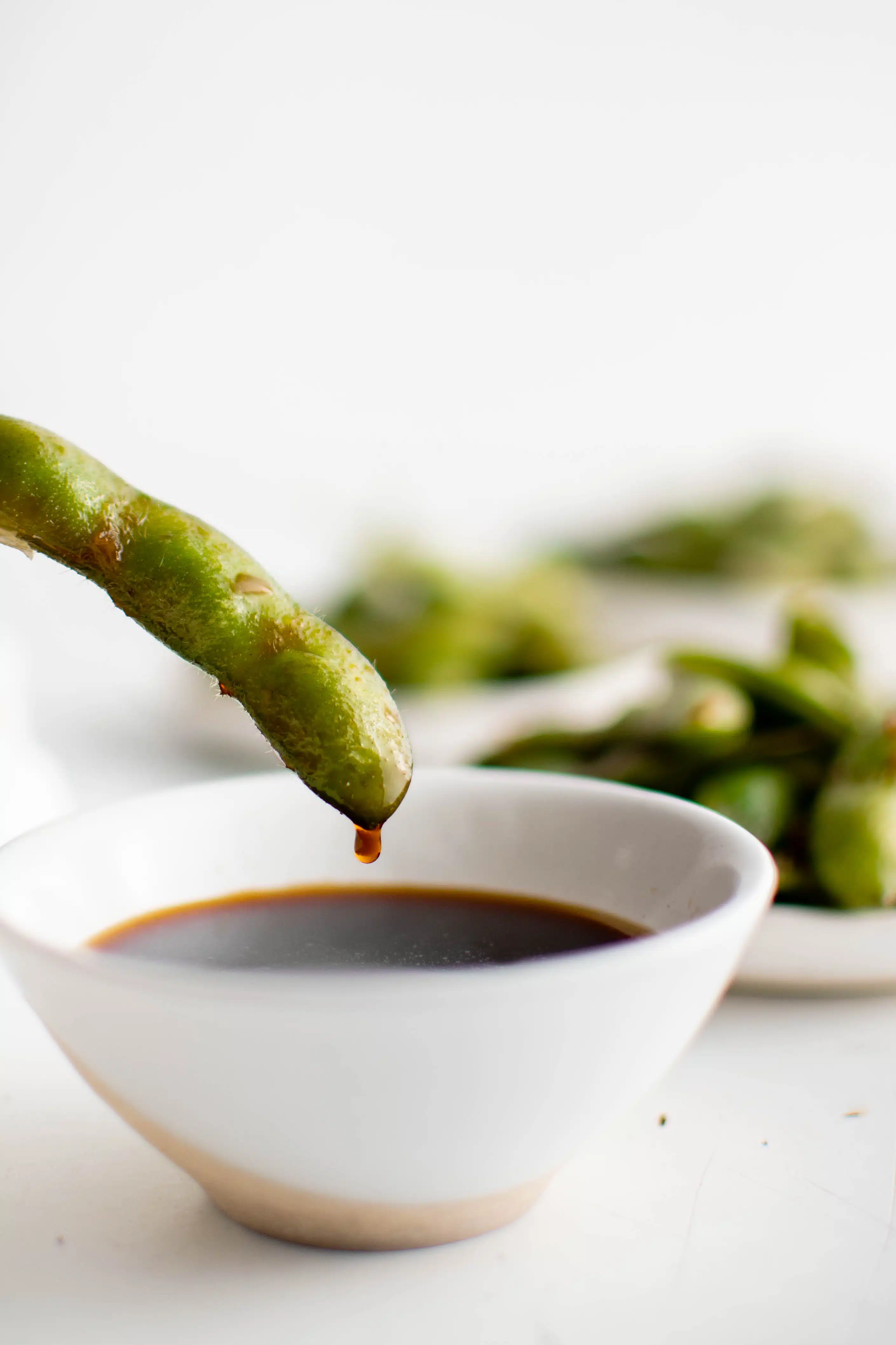 One soybean pod being dipping in a small bowl filled with soy sauce.
