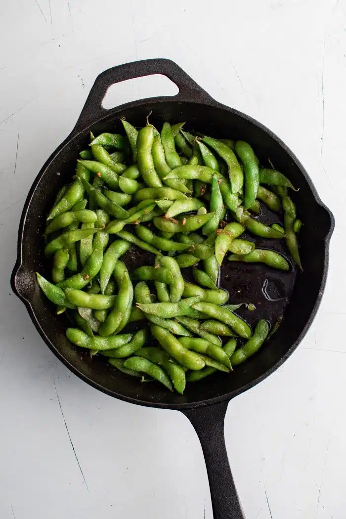 Large cast iron skillet filled with cooked edamame seasoned with soy sauce, garlic, sugar, and salt.