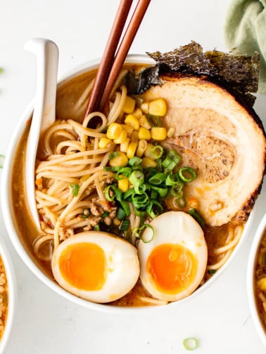 White bowl filled with miso ramen broth and ramen noodles topped with chashu pork, seaweed, ramen egg, corn, and green onions.