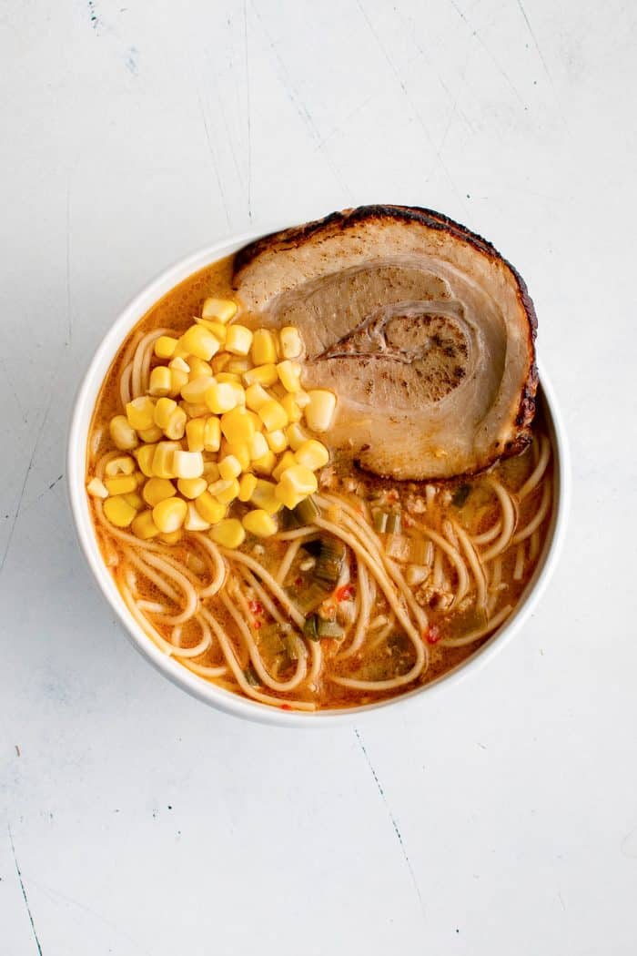 White bowl filled with miso ramen broth and ramen noodles topped with chashu pork, and sweet corn.