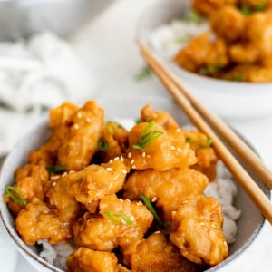 White bowl filled with white rice and topped with orange chicken pieces garnished with sliced green onions.