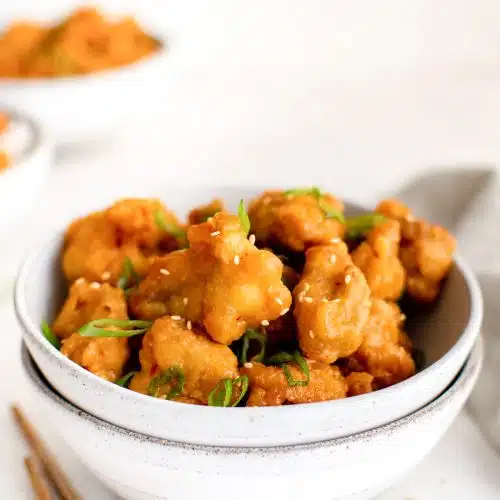 Stacked white bowls filled with orange chicken pieces garnished with sliced green onions.