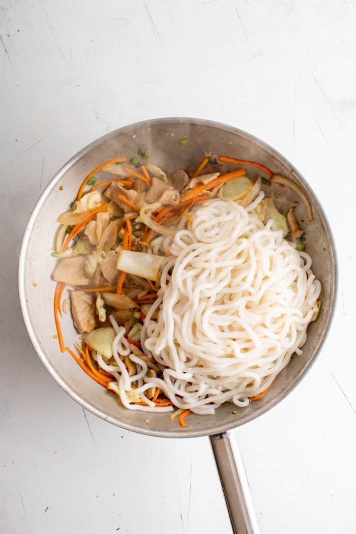 Udon noodles added to a large wok filled with stir-fried pork, vegetables, and sauce.