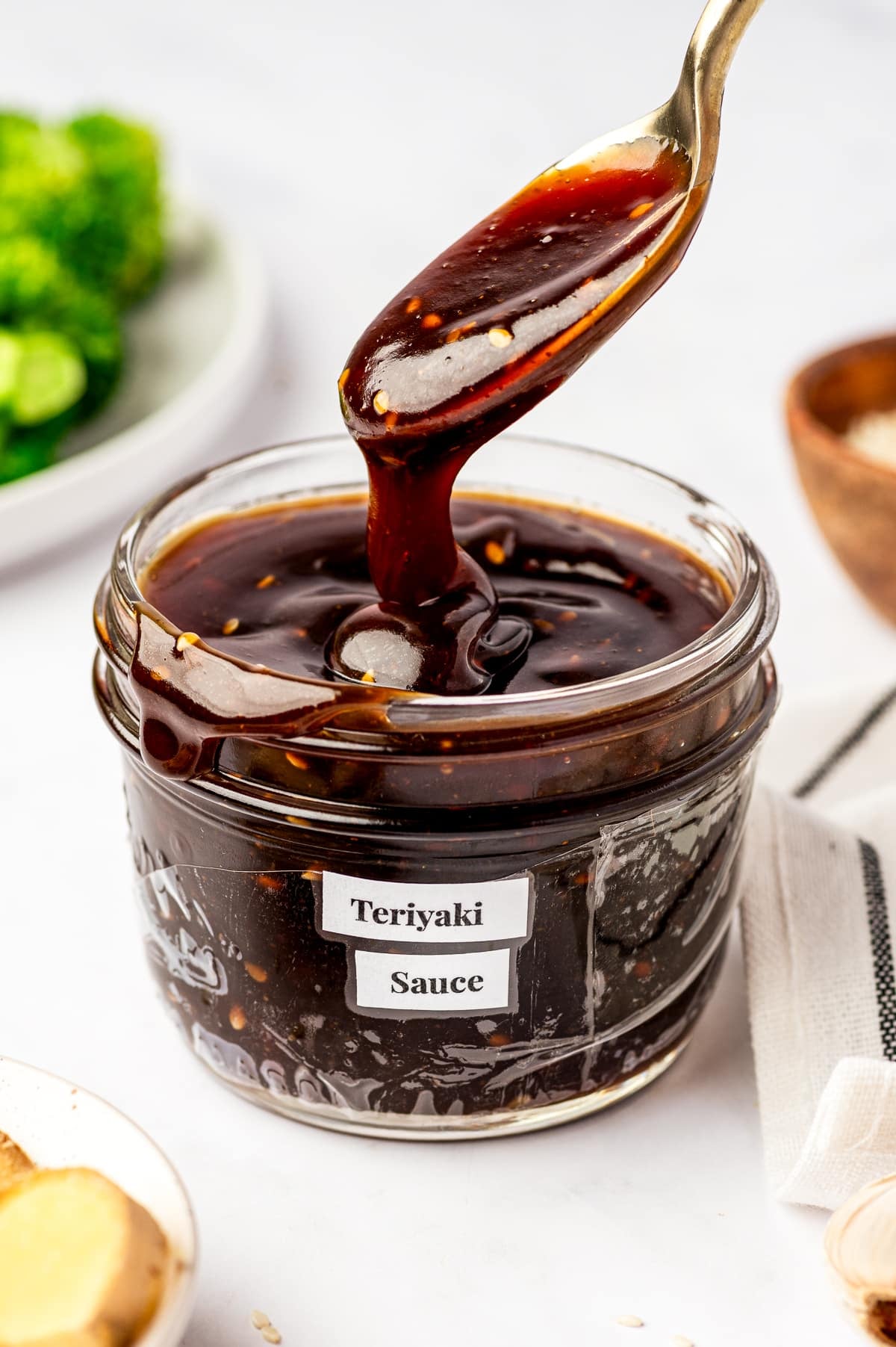 Thick, glossy teriyaki sauce dripping from a spoon back into a clear glass jar labeled "Teriyaki Sauce," with sesame seeds visible in the sauce and broccoli blurred in the background.