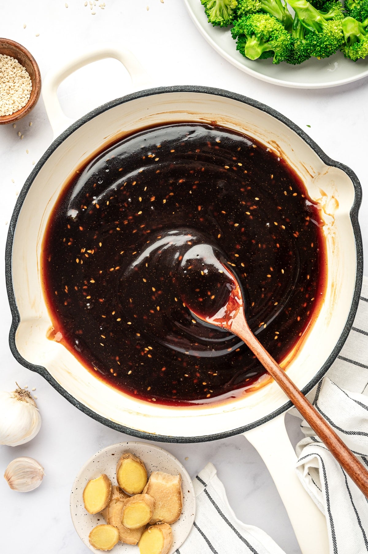 Thick homemade teriyaki sauce in a cream-colored pan with a wooden spoon, surrounded by broccoli, sesame seeds, garlic, fresh ginger slices, and a striped kitchen towel.