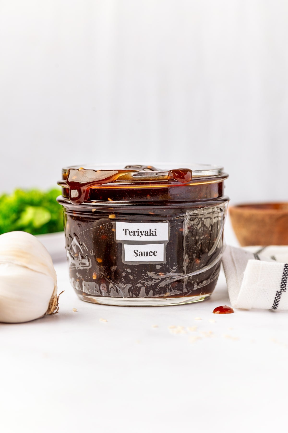 Clear glass jar filled with thick homemade teriyaki sauce and labeled "Teriyaki Sauce," with sauce on the rim, garlic on the side, sesame seeds scattered nearby, and a striped towel in the background.