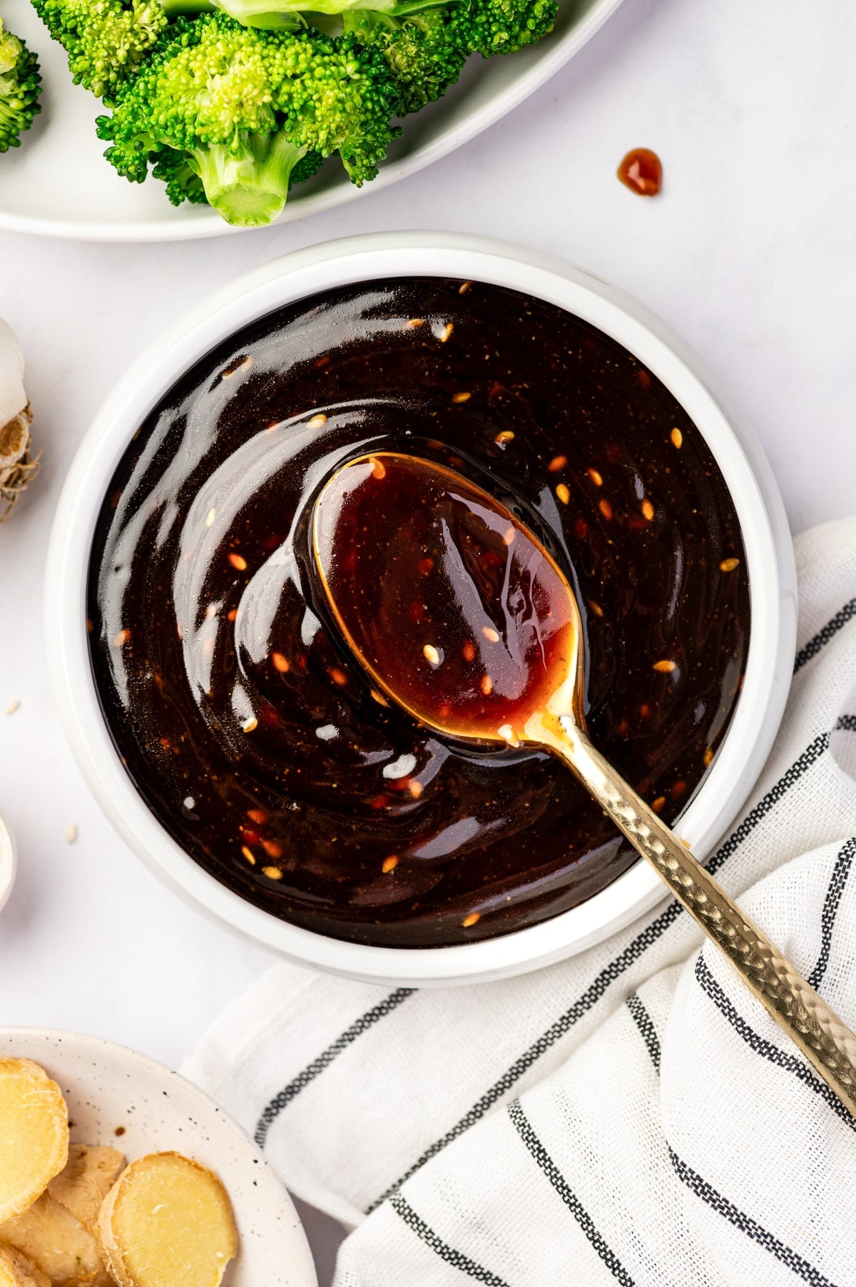 Homemade teriyaki sauce in a white bowl with a gold spoon, sesame seeds in the dark glossy sauce, broccoli at the top of the frame, fresh ginger slices, garlic, and a striped towel around the bowl.