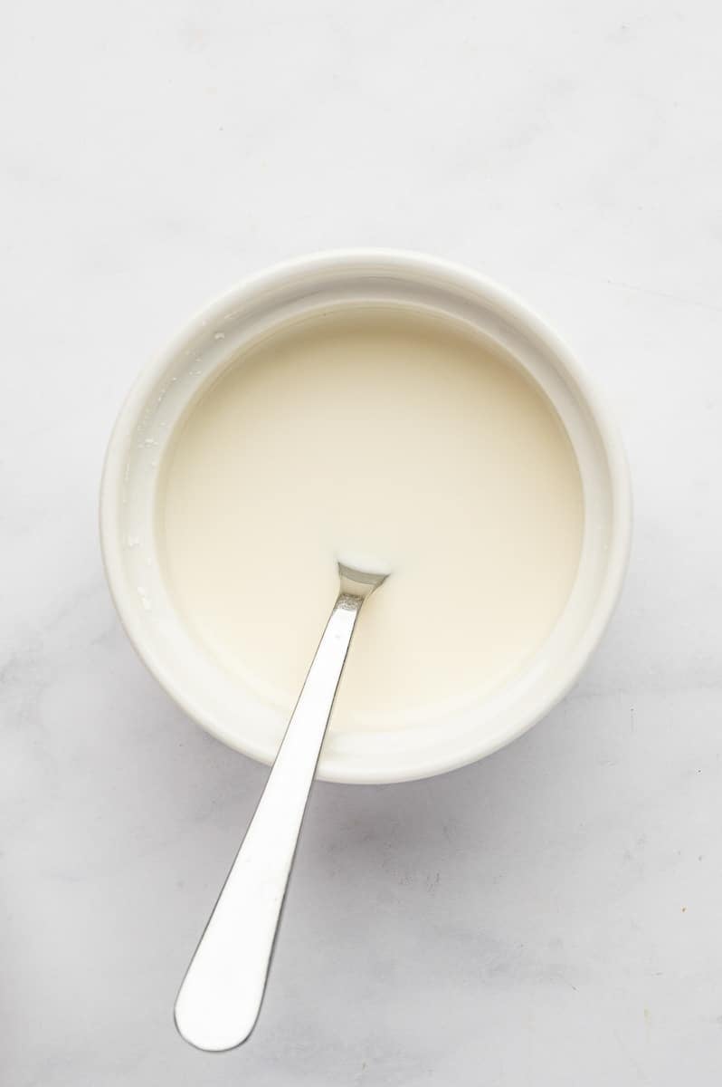 Cornstarch slurry mixed with cold water in a small white bowl with a silver spoon, shown on a light marble surface before being added to thicken teriyaki sauce.