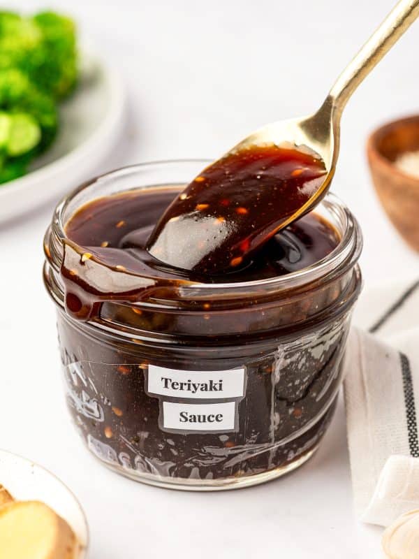 Thick homemade teriyaki sauce in a clear glass jar labeled "Teriyaki Sauce," with a spoon lifting the glossy dark sauce and broccoli, sesame seeds, garlic, and ginger in the background.