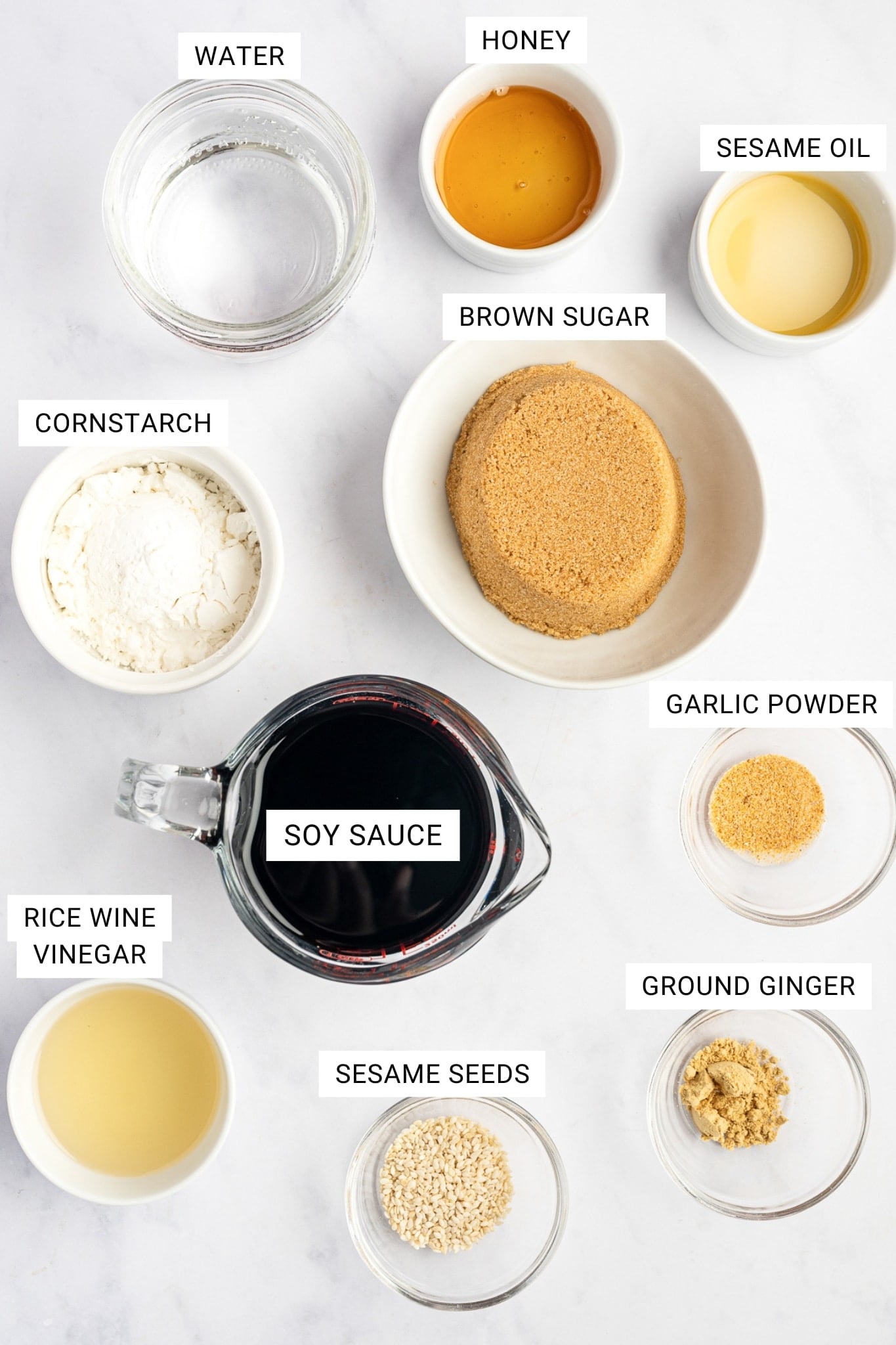 Labeled teriyaki sauce ingredients on a white marble surface, including water, honey, sesame oil, cornstarch, brown sugar, garlic powder, soy sauce, rice wine vinegar, sesame seeds, and ground ginger.