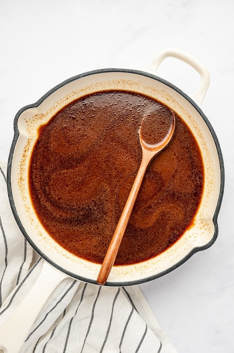 Homemade teriyaki sauce simmering in a cream-colored enamel pan with black trim, with a wooden spoon resting in the reddish-brown sauce and a striped kitchen towel beside the pan.