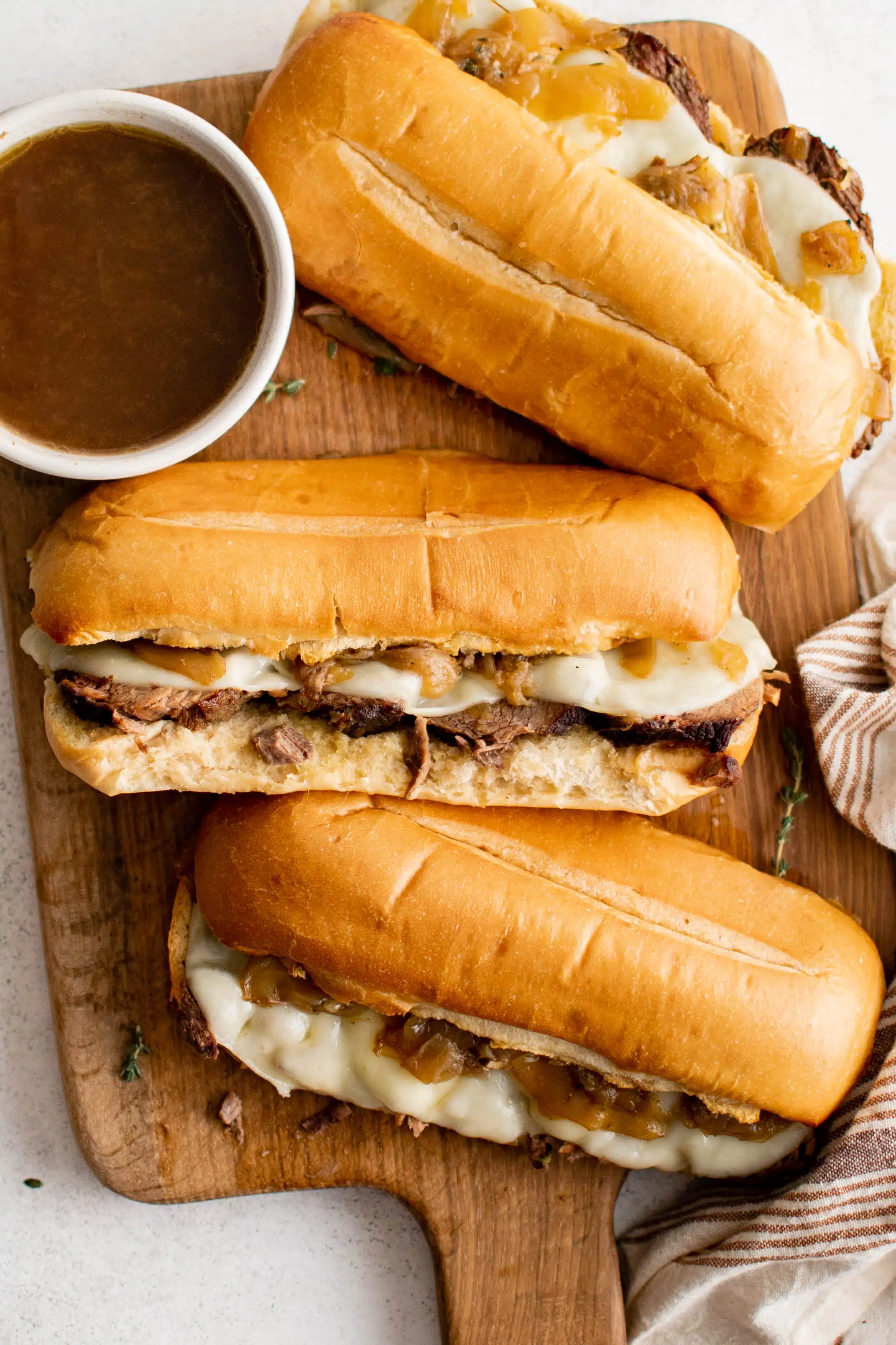 Three warm French dip sandwiches on a cutting board with a bowl of au jus ready to be enjoyed.