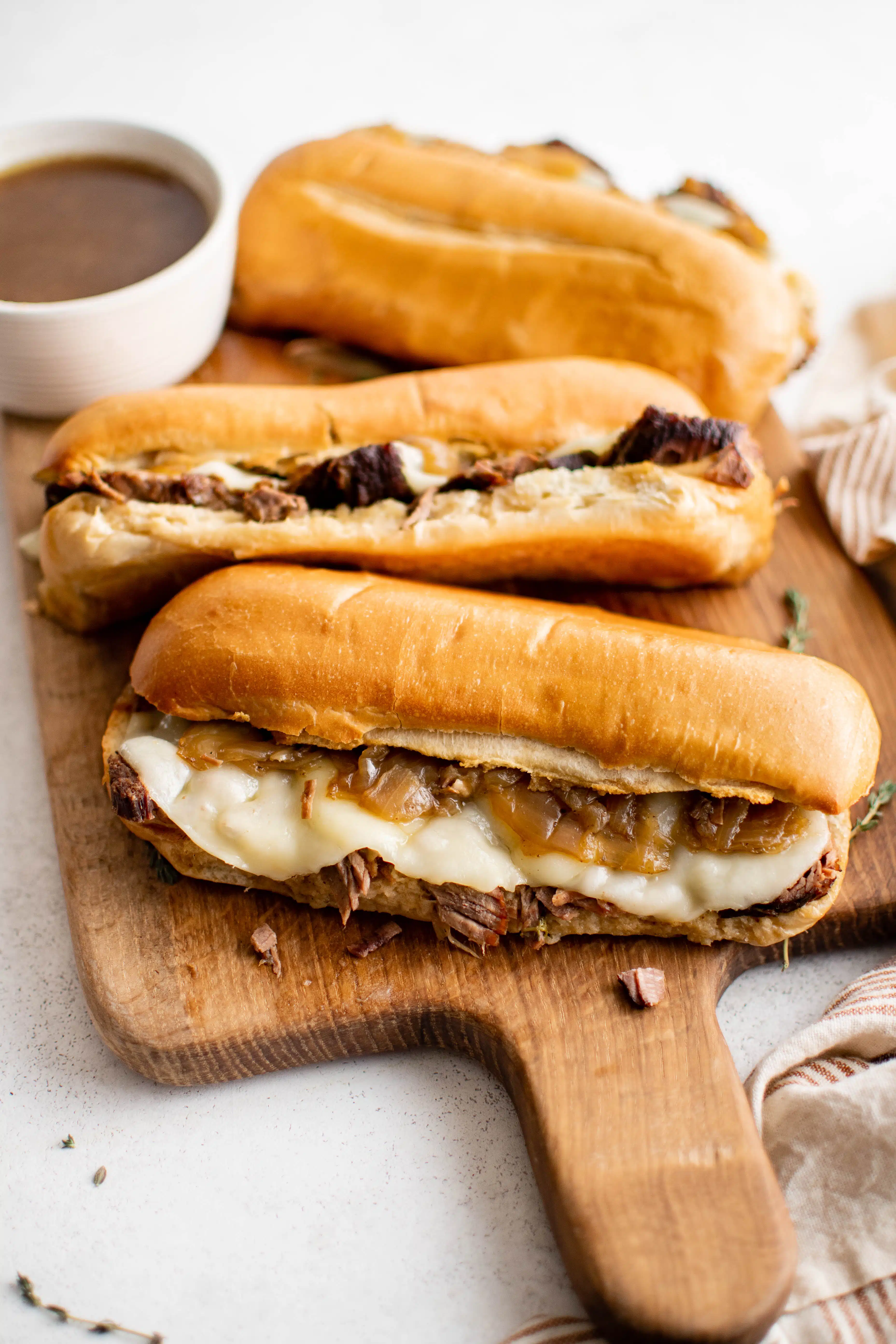 Three warm French dip sandwiches on a cutting board with a bowl of au jus ready to be enjoyed.