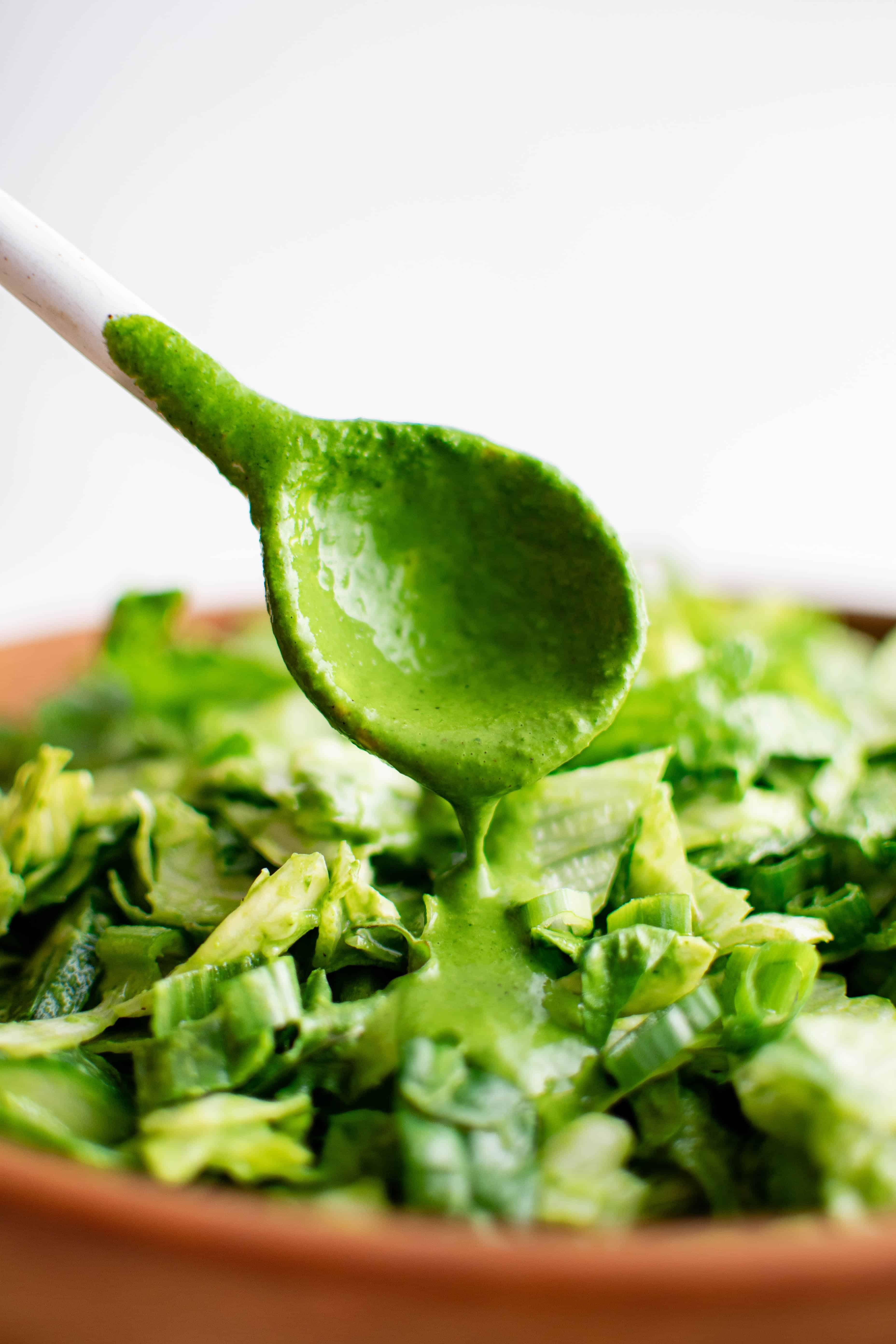 Wood spoon pouring homemade green goddess dressing over a bowl of chopped greens.