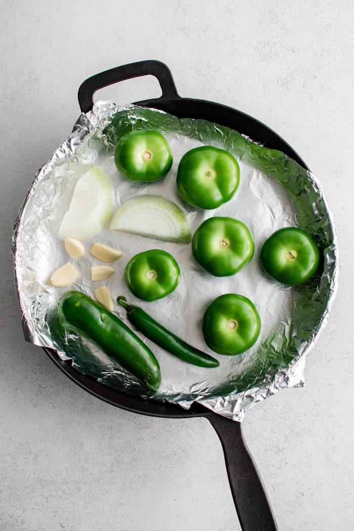 Whole raw tomatillos, jalapenos, garlic, and onion in a large cast iron pan lined with aluminum foil.