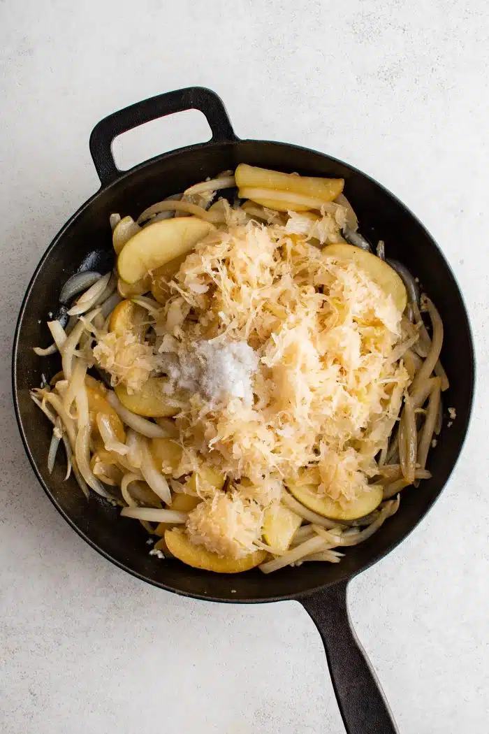 Salt and sauerkraut added to a large cast iron skillet filled with cooking onions and apples.
