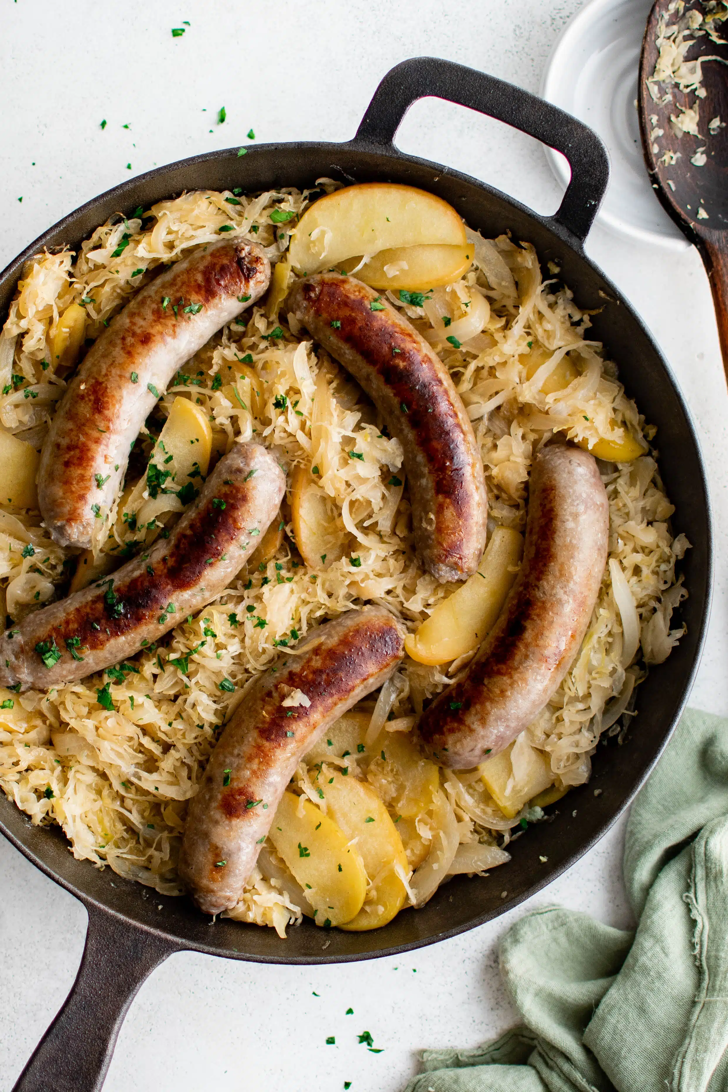 Five large pan-seared bratwurst nestled on top of a skillet filled with sauerkraut sauteed and mixed together with apples and onions and garnished with minced parsley.