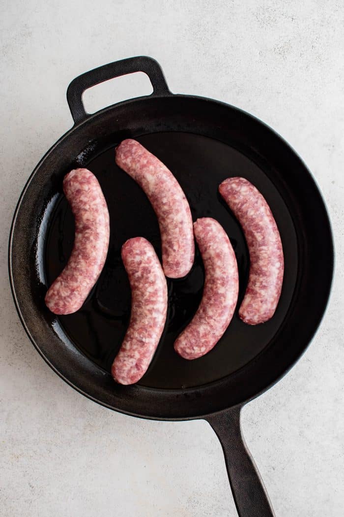 Large cast iron skillet with five raw cooking bratwurst.