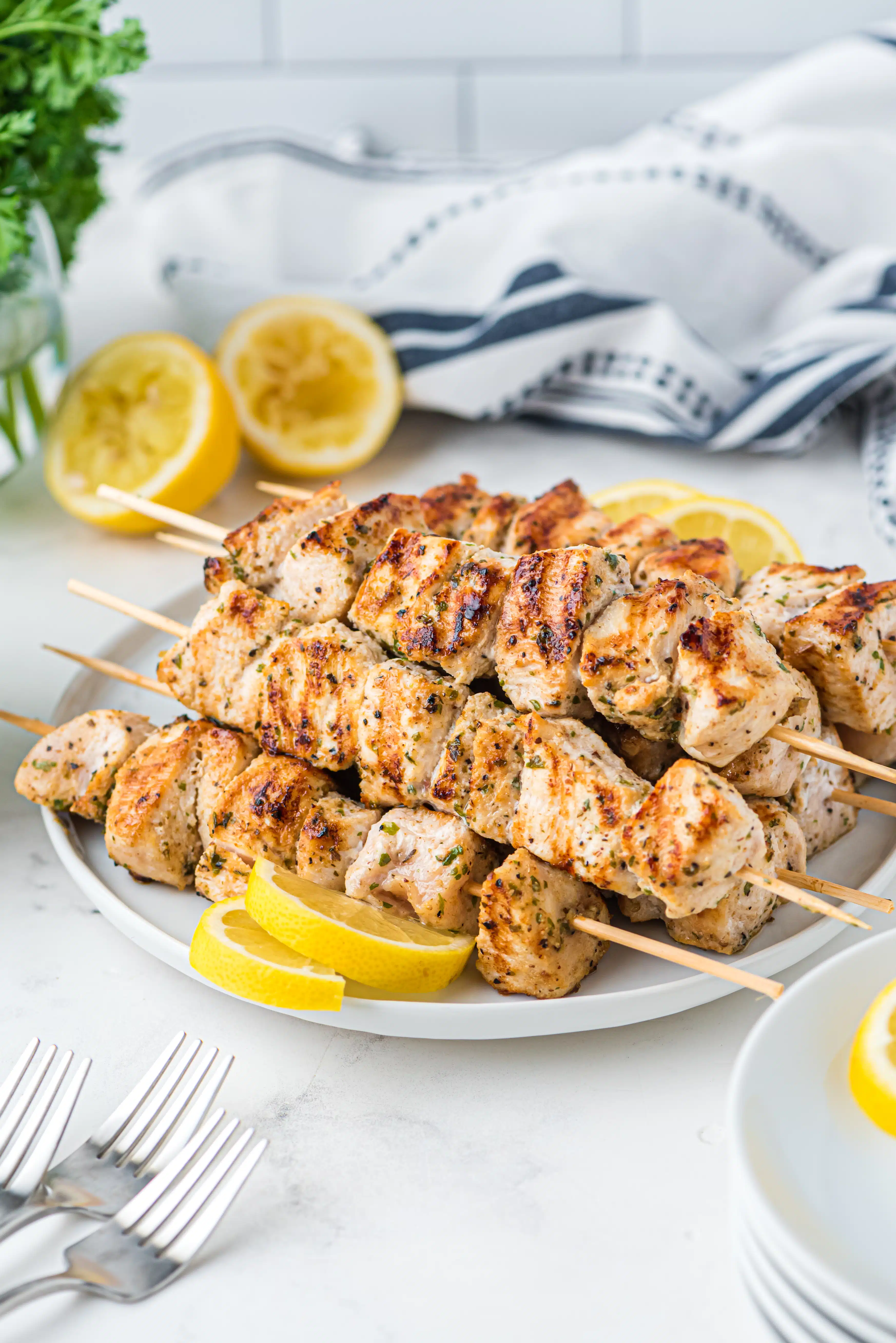 Pile of chicken skewers on a white plate with lemon slices and wedges for garnish.