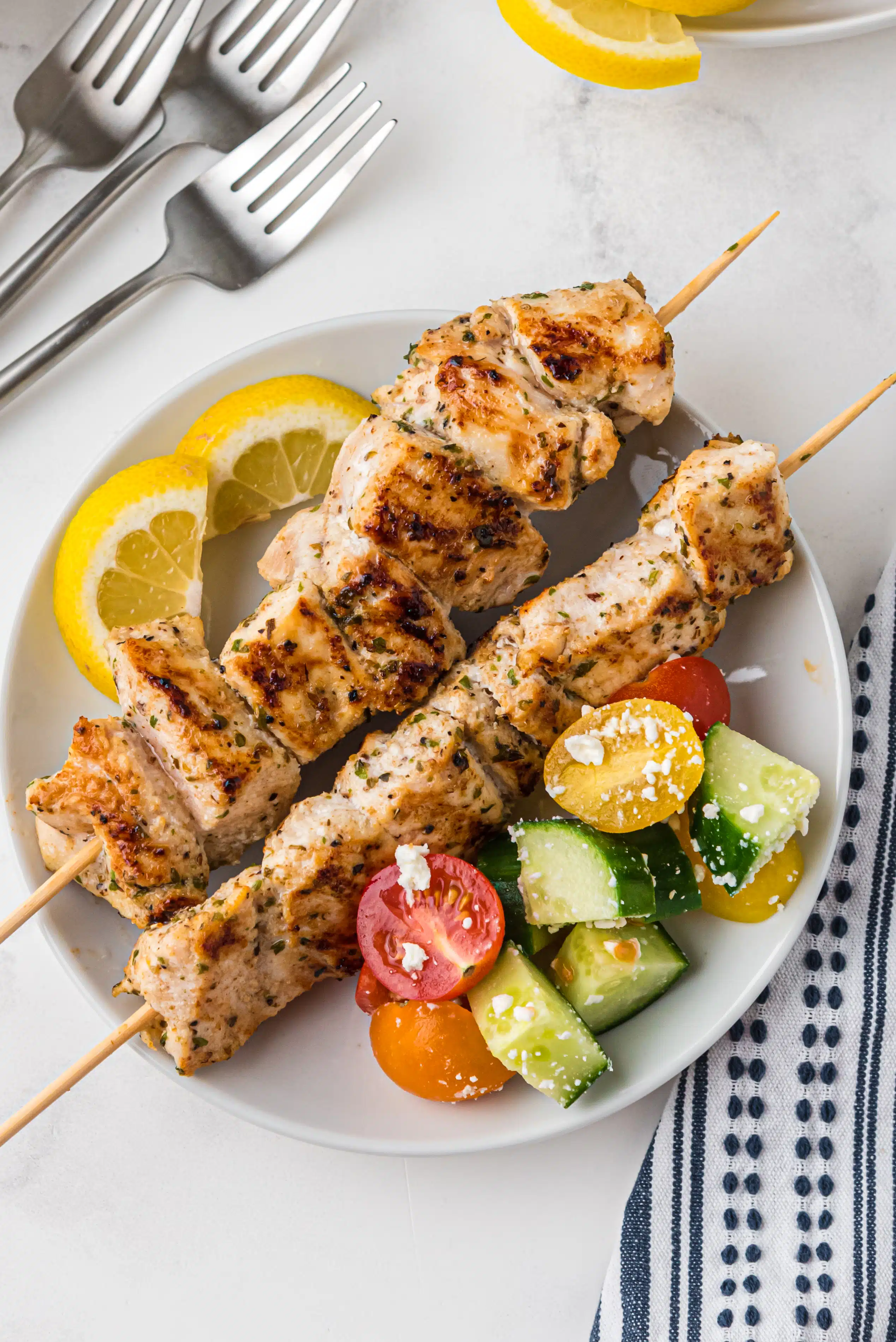 Dinner plate filled with two chicken skewers, lemon slices, and a side of greek salad.