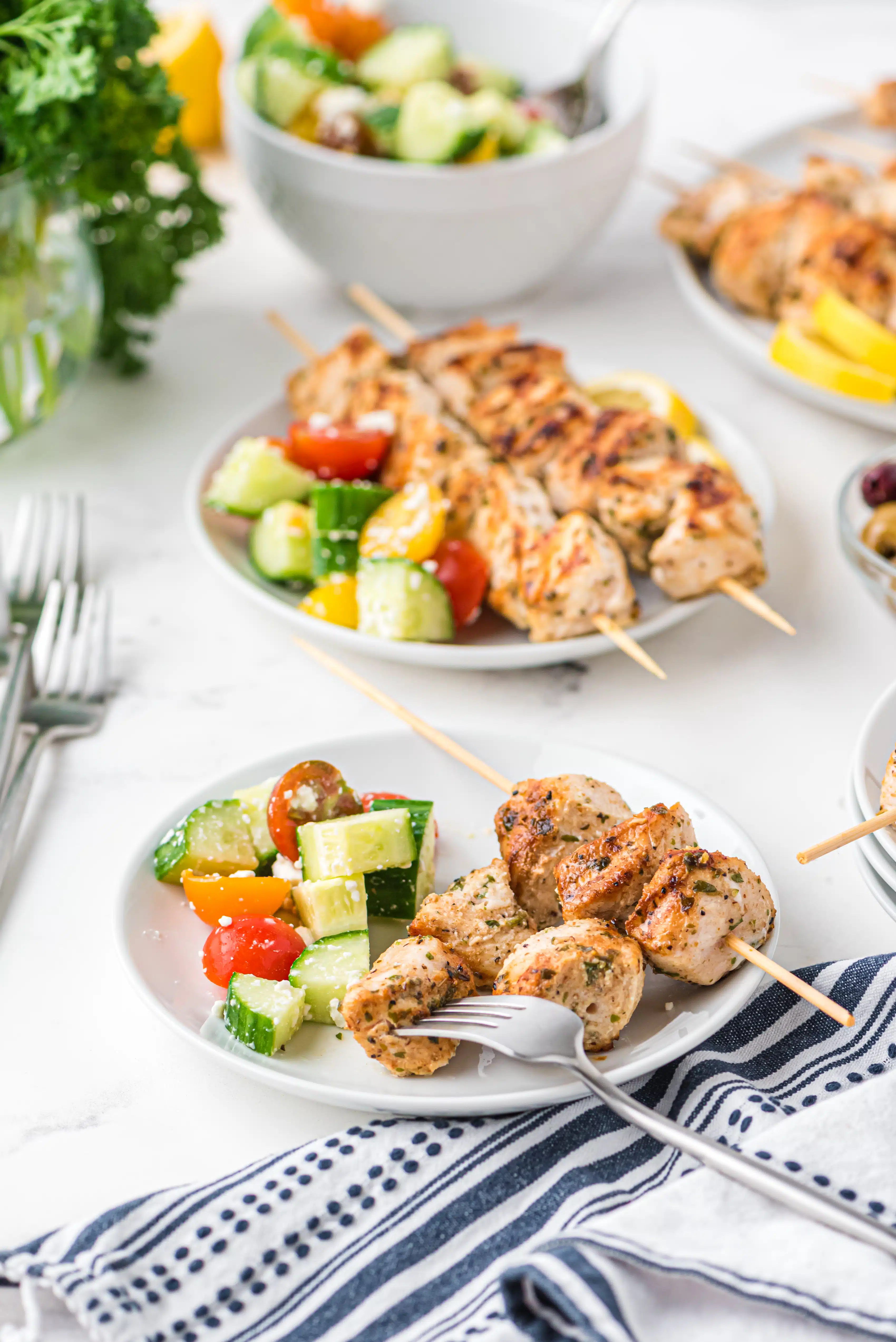 Two dinner plates filled with two chicken skewers, lemon slices, and a side of greek salad.