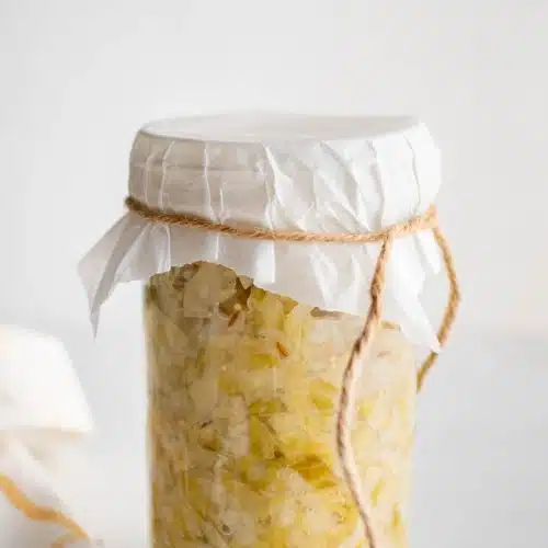 Tall glass jar filled with homemade sauerkraut with the top covered by white cloth and tied with string.