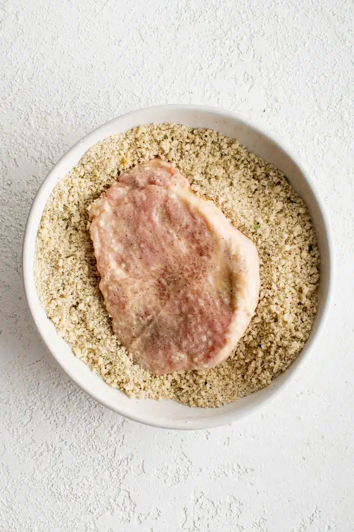 Pounded pork cutlet resting in a white bowl filled with seasoned panko breadcrumbs.