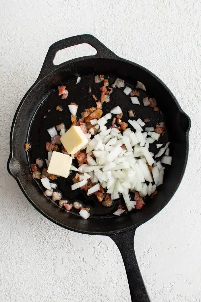 Two tablespoons of butter and diced onion added to a large cast iron skillet filled with cooking bacon pieces.