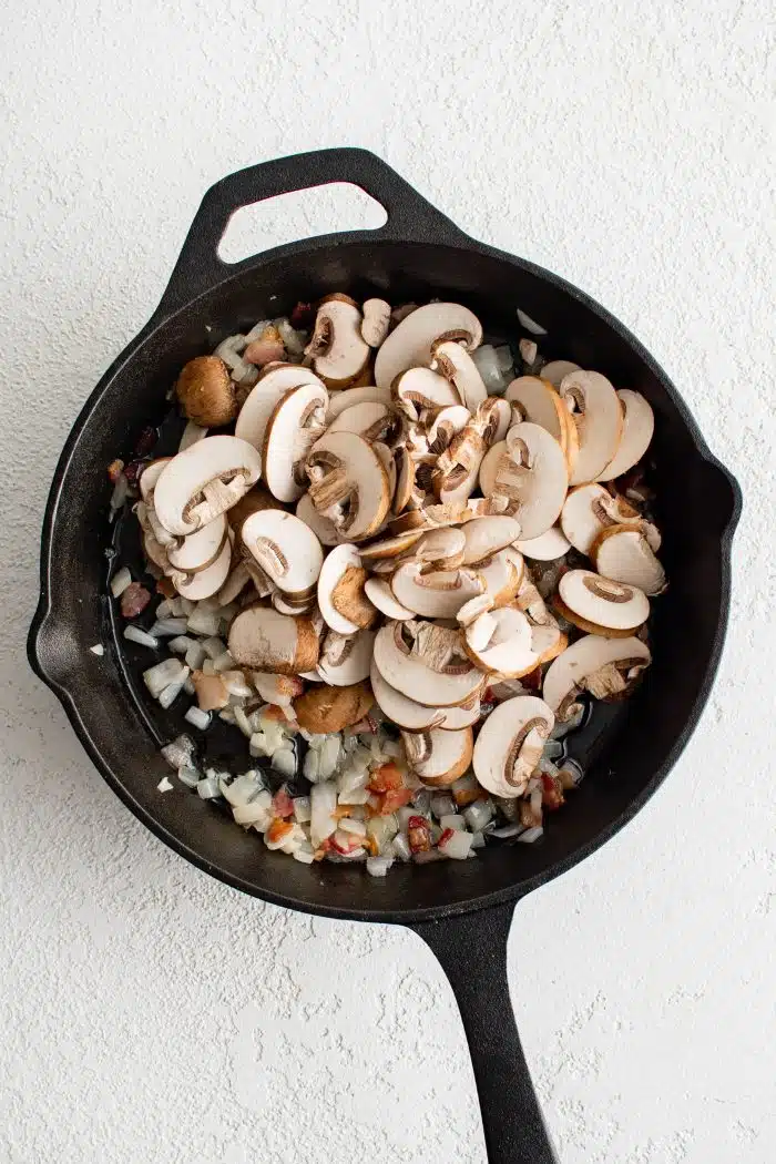Sliced cremini mushrooms added to the bacon and onions cooking in a large cast iron skillet.