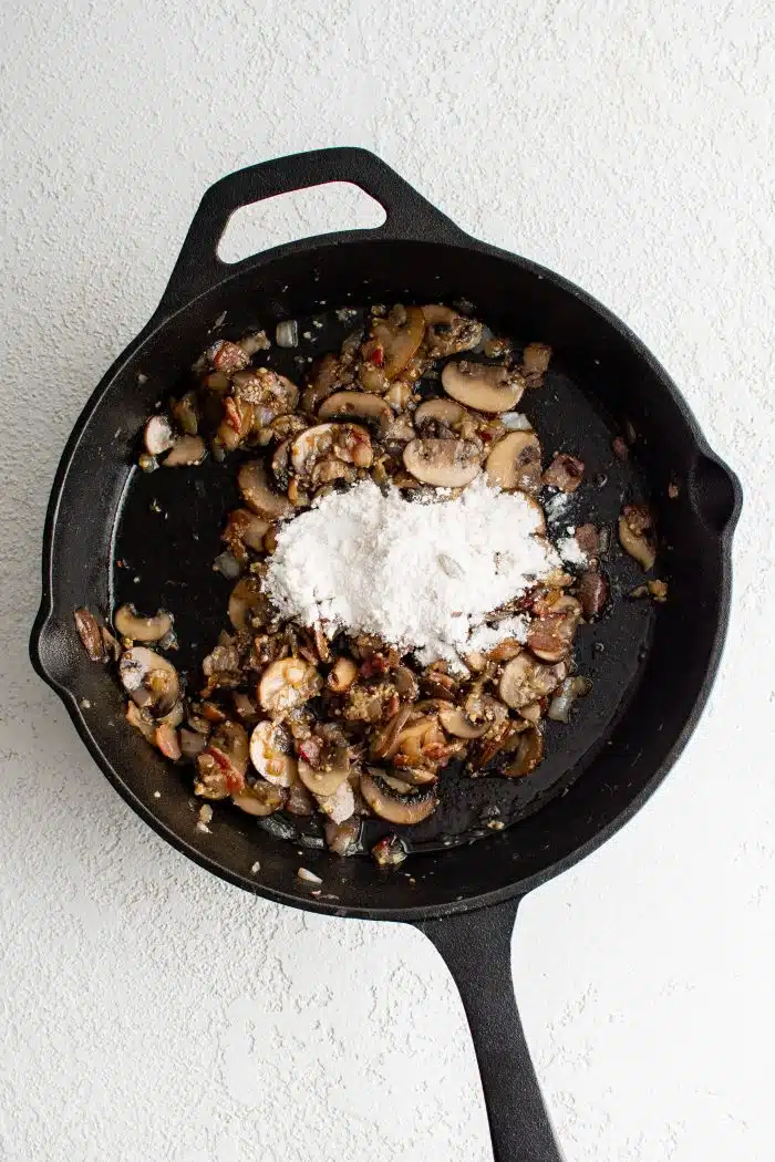 All-purpose flour added to mushrooms, onions, and bacon cooking in a large cast iron skillet.