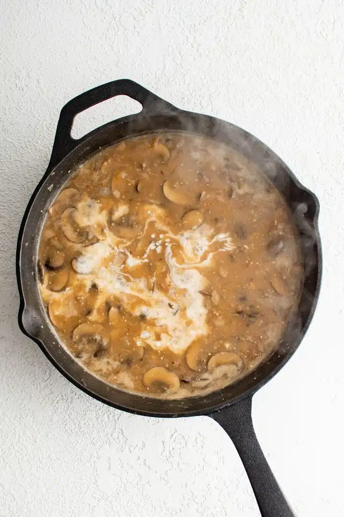 Half and half added to a large cast iron pan filled with a simmering sauce made with mushrooms and bacon.