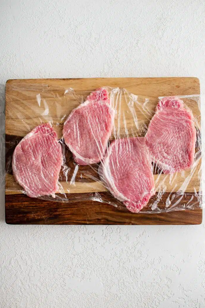 Four pork chops on a large cutting board and covered with plastic cling wrap.