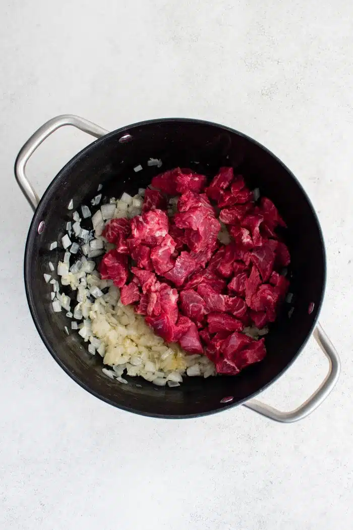 Chopped pieces of raw beef chuck in a large pot with cooking onions and minced garlic.