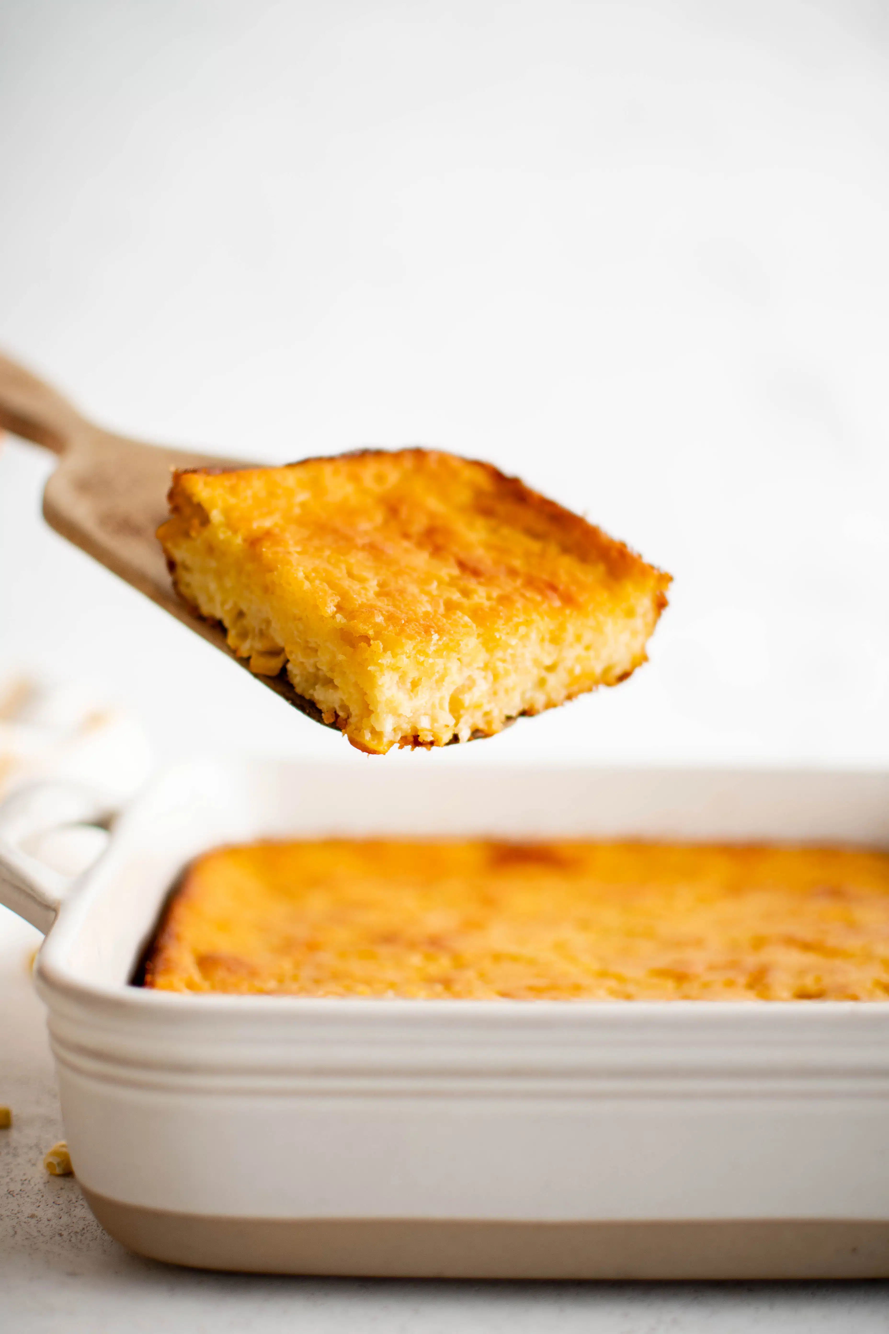 Wooden spatula holding a square of cooked cornbread casserole hovering above a white baking dish filled with golden brown baked cornbread casserole recipe.