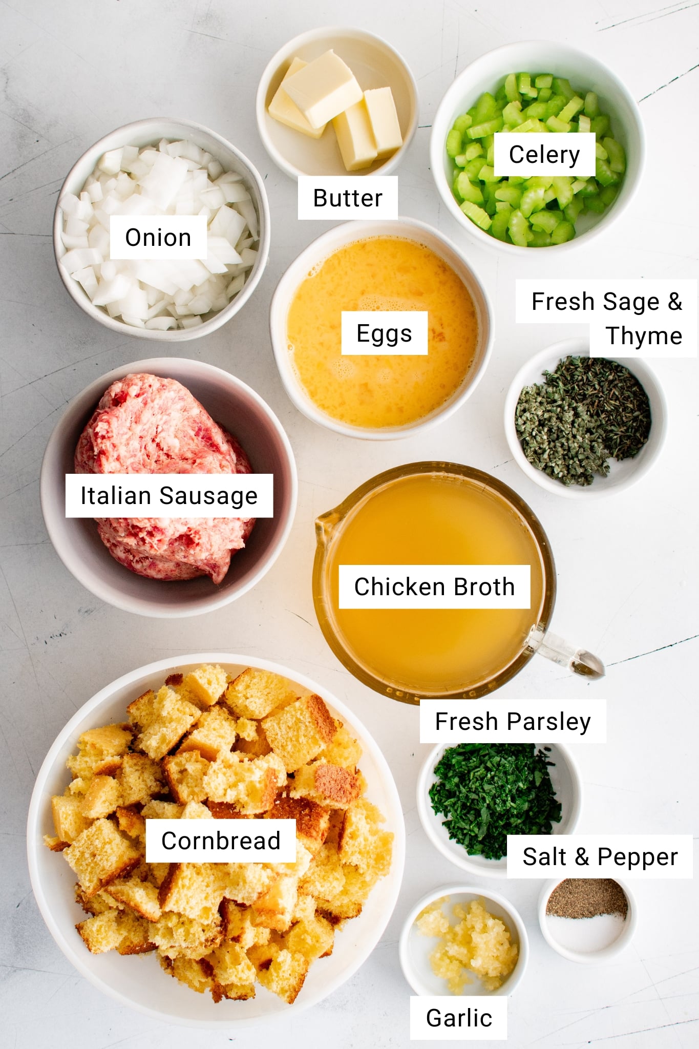 Overhead view of labeled ingredients for cornbread dressing including cubed cornbread, Italian sausage, onion, celery, chicken broth, eggs, butter, garlic, and fresh herbs.