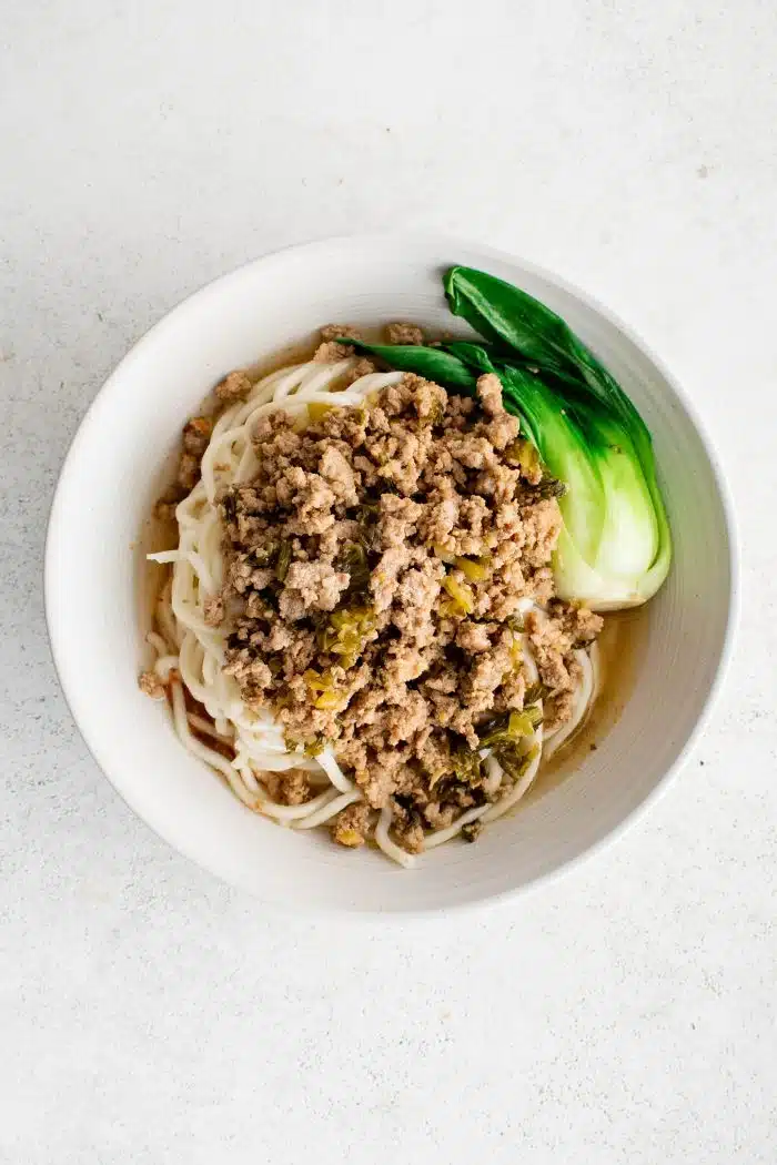 Large white bowl filled with spicy dan dan noodle sauce, cooked noodles, baby bok choy, cooked pork, and chicken broth.