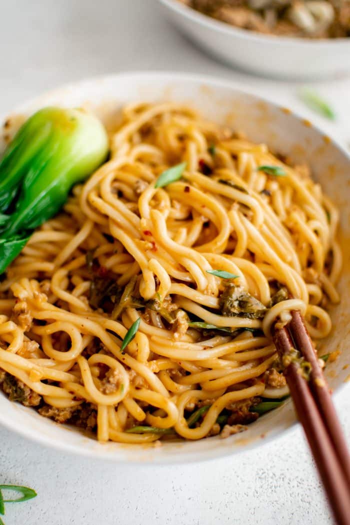 White bowl filled with cooked wheat noodles smothered in spicy sichuan sauce garnished with sliced green onions.