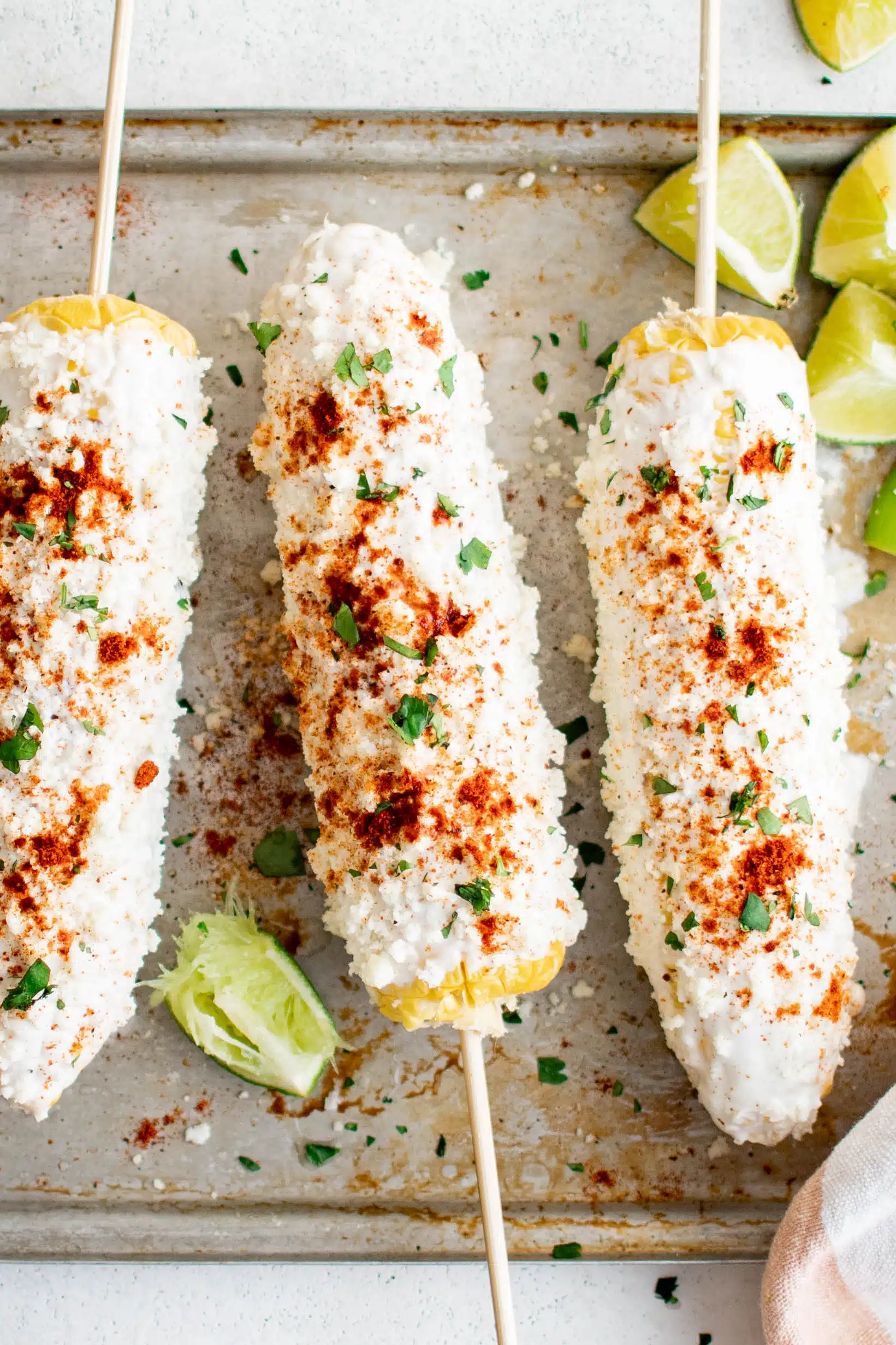 Three prepared elote on a baking sheet and garnished with paprika and chili powder, fresh cilantro, and lime wedges.