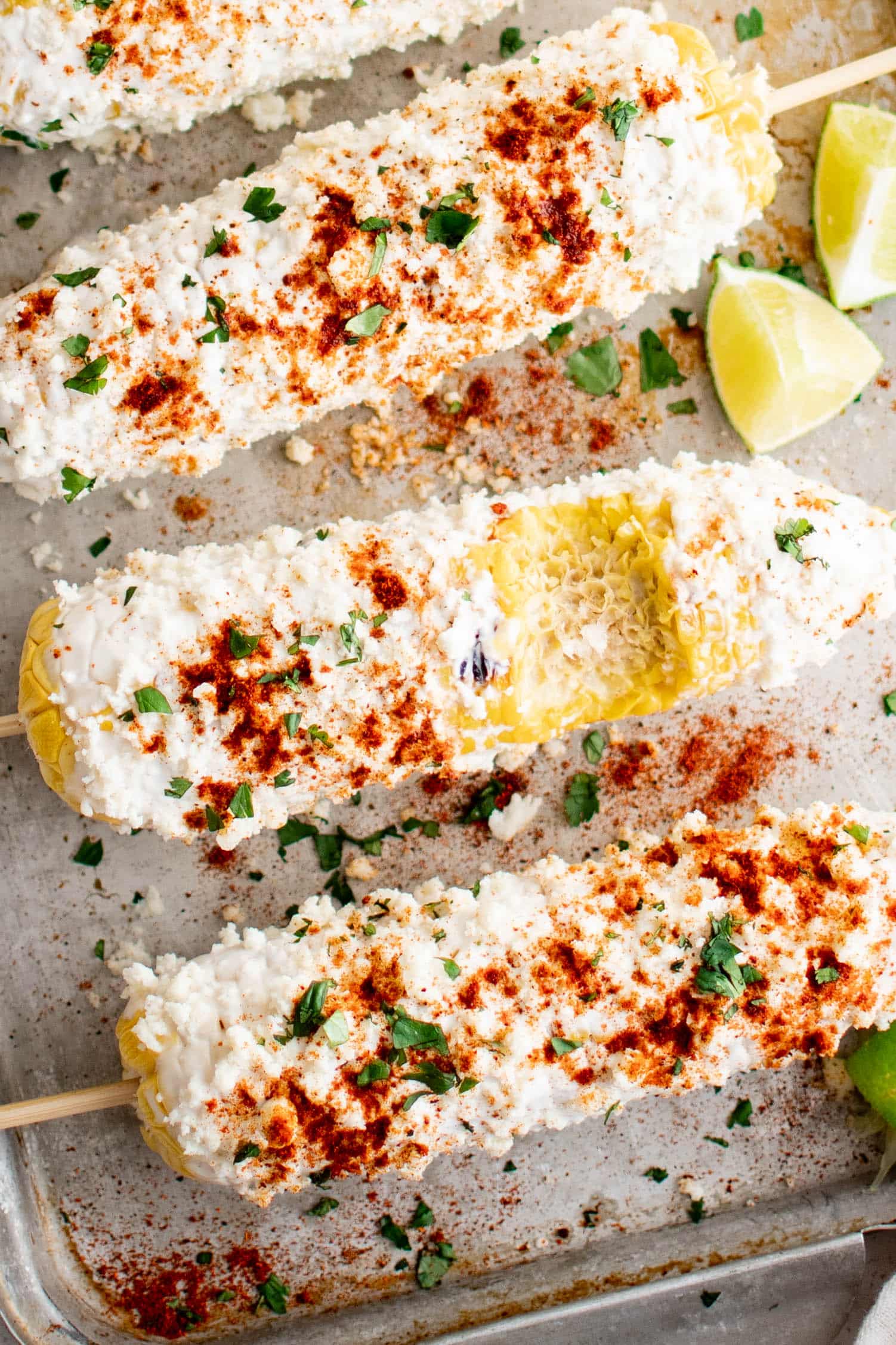 Three prepared elote - the middle with a bite taken out - on a baking sheet and garnished with paprika and chili powder, fresh cilantro, and lime wedges.