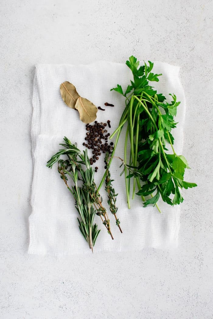 Fresh parsley, whole cloves, fresh rosemary, bay leaves, and black pepper corns set on top of white cheesecloth that will be used as an herb bag.