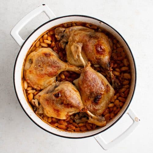 Overhead image of a large white Dutch oven filled with a bottom layer of chopped pork belly and sausage, a middle layer of white beans, and a top layer of four crispy duck legs.