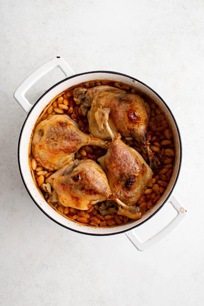 Overhead image of a large white Dutch oven filled with a bottom layer of chopped pork belly and sausage, a middle layer of white beans, and a top layer of four crispy duck legs.