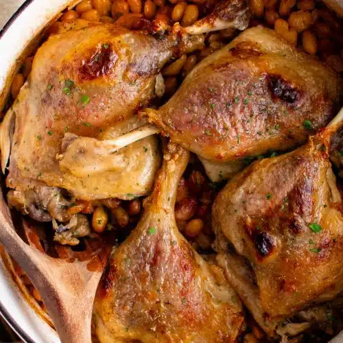 Overhead image of a large white Dutch oven filled with a bottom layer of chopped pork belly and sausage, a middle layer of white beans, and a top layer of four crispy duck legs.
