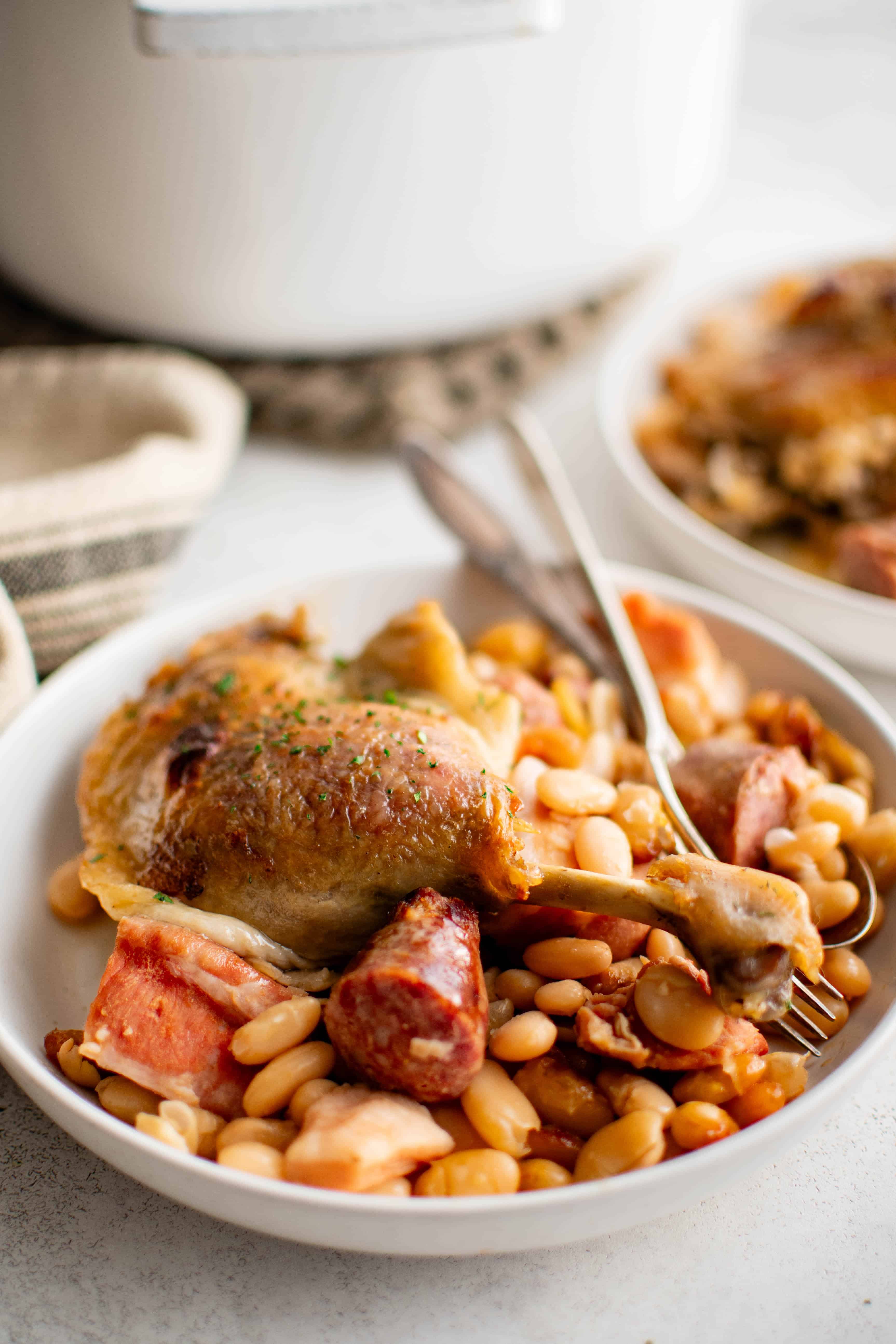 White dinner plate filled with a serving of cassoulet consisting of sausage, pork belly, beans, and duck leg.