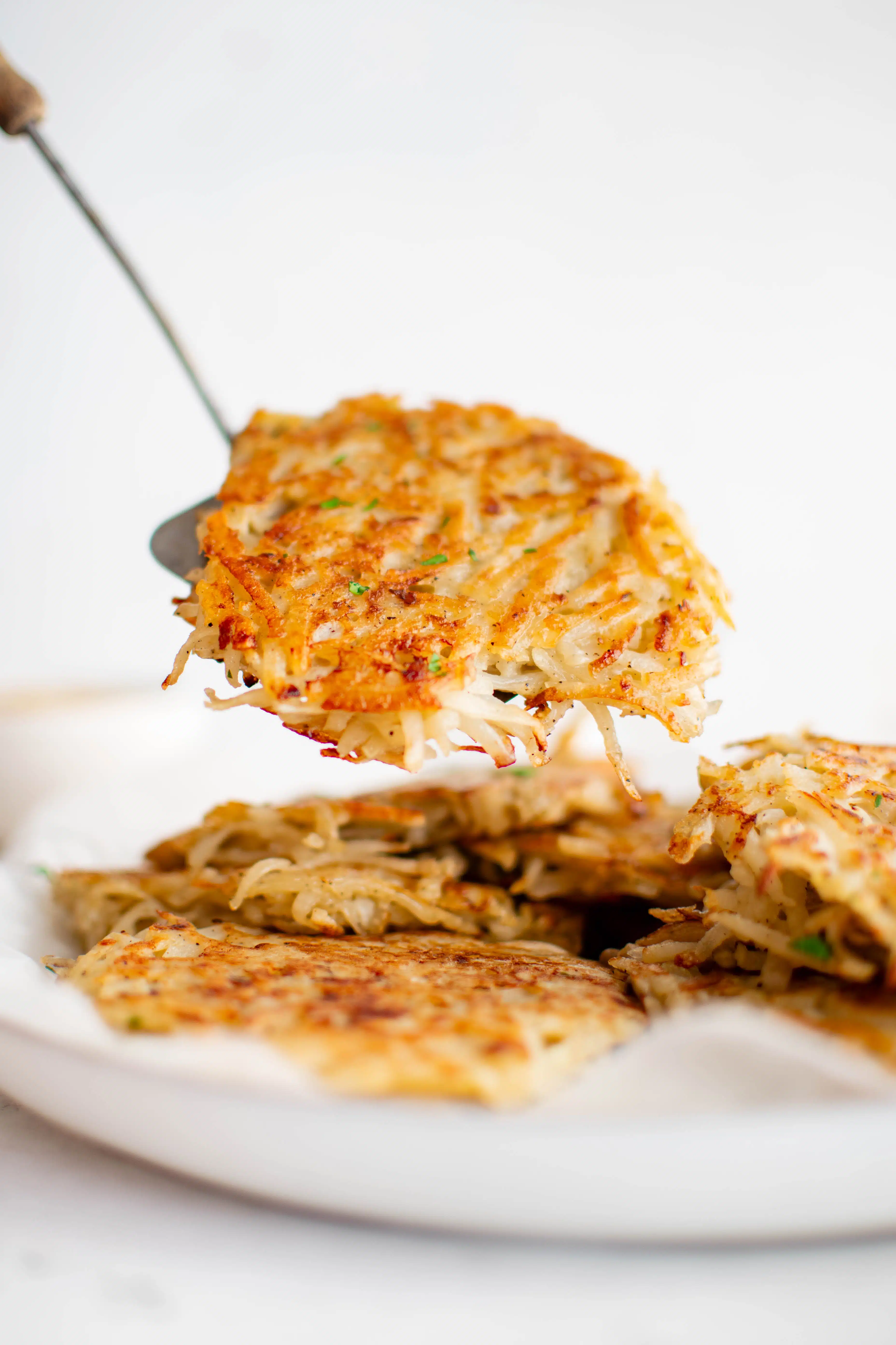 Metal spatula holding a single German potato pancake hovering over a plate filled with more potato pancakes.